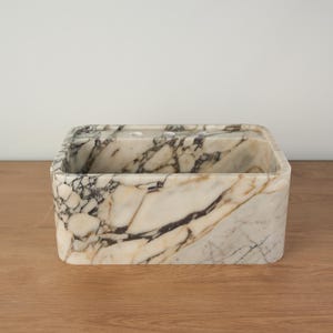 May include: A rectangular marble sink with rounded corners. The sink features a cream-colored base with dark gray and brown veining throughout. The sink has a small hole on the top surface. The sink is sitting on a wooden surface.