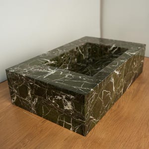 May include: A rectangular, dark green marble sink with white veining. The sink has a recessed basin and is set on a wooden surface. The marble has a polished finish, showcasing the natural patterns.