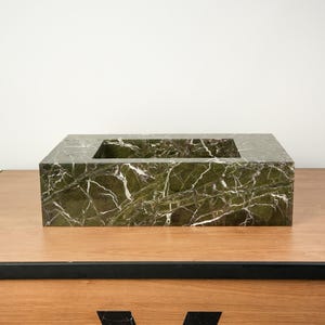 May include: A rectangular, green marble container with a square opening in the center. The marble has white veining throughout. The container is displayed on a wooden surface.