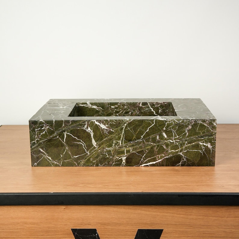 May include: A rectangular, green marble container with a square opening in the center. The marble has white veining throughout. The container is displayed on a wooden surface.