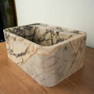May include: A rectangular marble container with rounded corners. The container features a cream-colored base with dark gray and brown veining. The interior has a divider, and the marble has a polished finish. The container sits on a wooden surface.