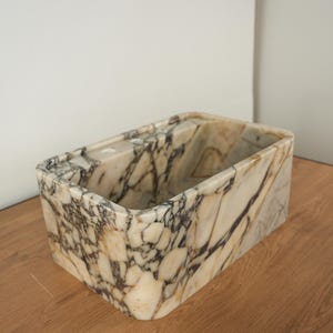 May include: Rectangular marble container with rounded corners. The container features a cream-colored base with dark gray and brown veining throughout. The interior is hollow, and the container sits on a wooden surface.