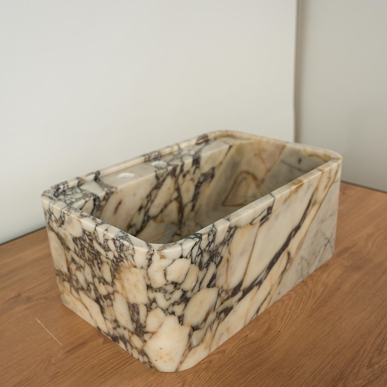 May include: Rectangular marble container with rounded corners. The container features a cream-colored base with dark gray and brown veining throughout. The interior is hollow, and the container sits on a wooden surface.