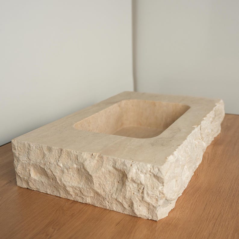 Pu&ograve; includere: Un lavandino rettangolare in pietra beige con un bacino scavato. Il lavandino ha un esterno ruvido e strutturato e poggia su una superficie di legno. Il bacino interno &egrave; liscio e il design generale &egrave; minimalista.