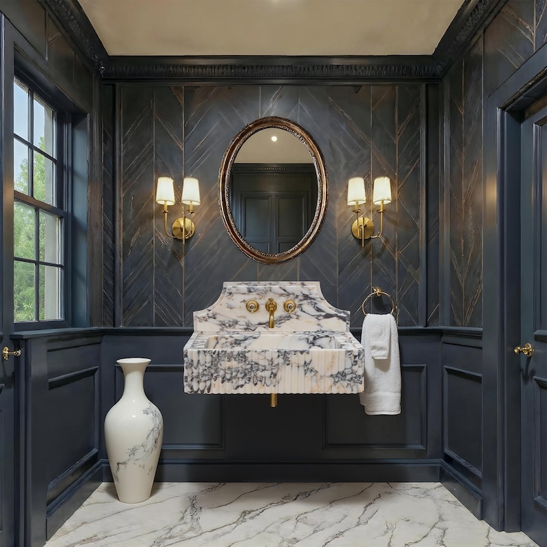 May include: A luxurious bathroom with a marble sink, gold fixtures, and a decorative oval mirror. The walls are dark blue with a herringbone pattern, and the floor is marble. A white vase stands in the corner, and a white towel hangs on a gold ring.