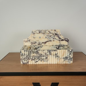 May include: A decorative marble box with a lid. The box is cream-colored with dark purple veining. The front of the box has vertical ridges. The lid has a curved top with three small circular holes. The box is sitting on a wooden surface.