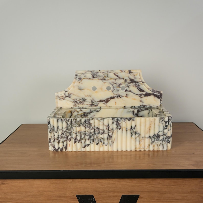 May include: A decorative marble box with a lid. The box is cream-colored with dark purple veining. The front of the box has vertical ridges. The lid has a curved top with three small circular holes. The box is sitting on a wooden surface.