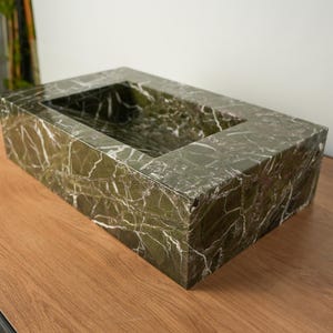 May include: A rectangular, green marble sink with white veining. The sink has a recessed basin and is displayed on a wooden surface. The natural stone has a polished finish, showcasing the intricate patterns of the marble.