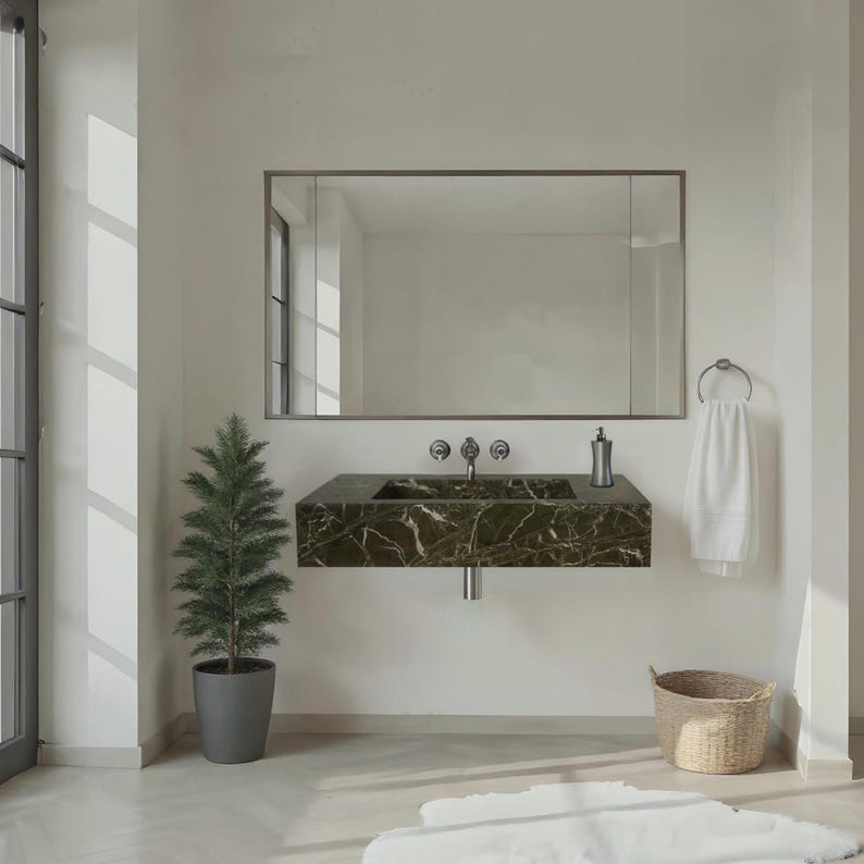 May include: A modern bathroom scene featuring a dark green marble sink with chrome fixtures. A large mirror hangs above the sink. A small potted evergreen tree and a woven basket add natural elements to the space. A white towel hangs on a chrome ring.