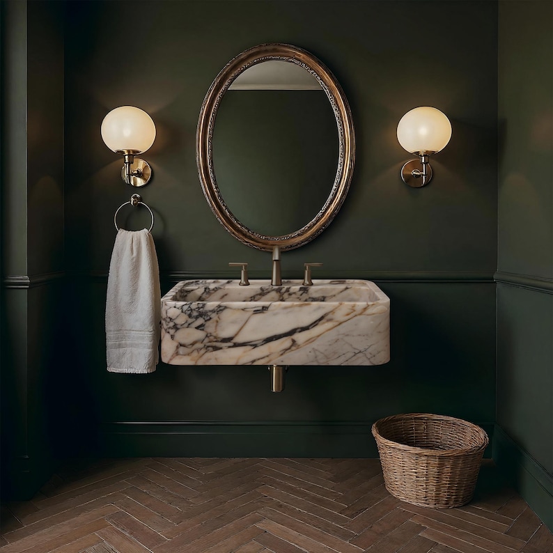 May include: A bathroom interior featuring a marble sink with brass fixtures, an oval mirror, and two globe-shaped wall sconces. A white towel hangs on a brass ring. A woven basket sits on the herringbone-patterned wood floor. The walls are painted a dark green.