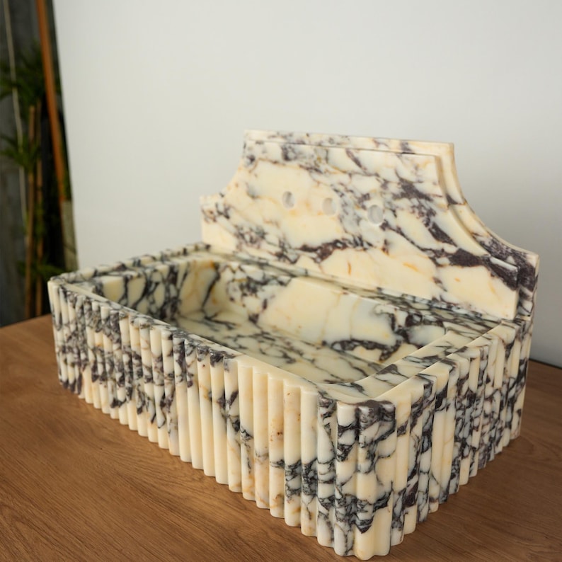 May include: A rectangular marble sink with a cream and dark brown veined pattern. The sink has a fluted exterior and a decorative back with three holes. The sink is set on a wooden surface.