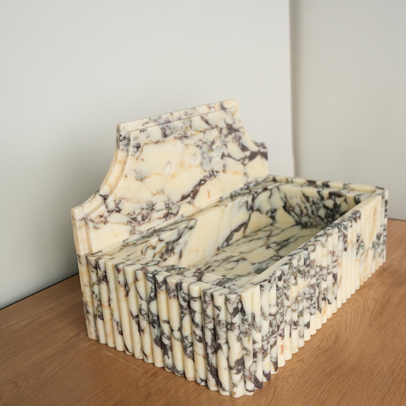 May include: A rectangular marble sink with a decorative back. The sink features a cream and brown marbled pattern. The front and sides have a vertical ribbed design. The sink is placed on a wooden surface.
