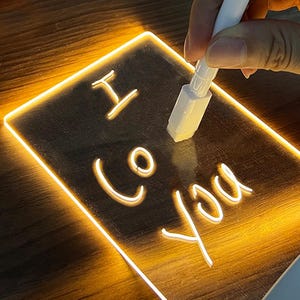 May include: A rectangular LED message board, illuminated with a warm yellow light, displaying the words "I Co You" in white. A white pen is held near the board, ready to write. The board is on a wooden surface.