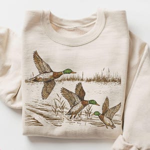 May include: Cream-colored sweatshirt featuring a detailed illustration of three ducks in flight over water. The ducks have brown and white wings and green heads. The sweatshirt has a crew neck and long sleeves. The words "NEXT DAY SHIPPING" are in a red circle.