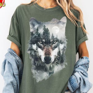 May include: Olive green t-shirt featuring a detailed wolf graphic with a forest and moon design. The wolf's face is the focal point, with intricate fur details and piercing eyes. The shirt has a relaxed fit and rolled sleeves.