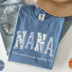 May include: A light blue t-shirt with the word "NANA" in a patchwork design. The letters are decorated with floral and gingham patterns. Below the word are the names "bella maria angel zoe". A pair of blue jeans and a tan hat are in the background.