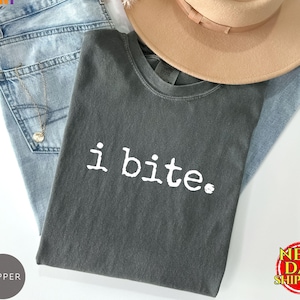 May include: A dark gray t-shirt with the white text "i bite." The shirt is folded on top of a pair of light blue jeans. A tan hat and a gold necklace are also in the image.