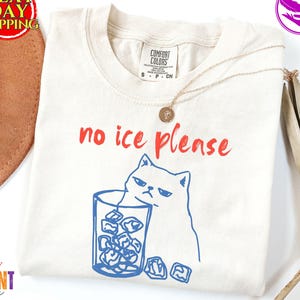 May include: Cream-colored t-shirt with the phrase "no ice please" in red script. A blue line drawing of a grumpy cat is next to a glass of ice cubes. The shirt has a Comfort Colors label.