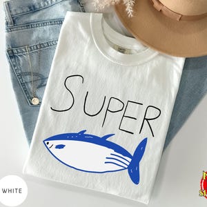May include: White t-shirt with the word "SUPER" in black and a blue and white fish graphic. The shirt is laid on top of blue jeans, with a tan hat and a gold necklace visible.
