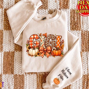 May include: A beige crewneck sweatshirt featuring the word GIGI in large, patterned letters with orange plaid, floral, leopard print, and polka dot designs. The graphic includes pumpkins, sunflowers, and a bow. The sleeve displays the names Olivia, Melissa, and Peter with a small pumpkin icon.
