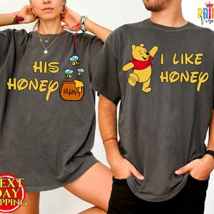 May include: Two charcoal gray t-shirts with Winnie the Pooh designs. One shirt says "HIS HONEY" with bees and a honey pot graphic. The other says "I LIKE HONEY" with a Pooh Bear graphic. The shirts are part of a matching set.