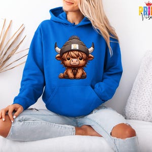Op de afbeelding: Een koningsblauwe hoodie met een cartoon illustratie van een Schotse Hooglander die een muts draagt. De koe is bruin met zwarte hoorns en een geruit overhemd. De hoodie heeft een voorzak en een capuchon met trekkoord.