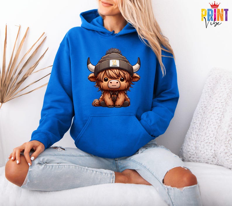 Op de afbeelding: Een koningsblauwe hoodie met een cartoon illustratie van een Schotse Hooglander die een muts draagt. De koe is bruin met zwarte hoorns en een geruit overhemd. De hoodie heeft een voorzak en een capuchon met trekkoord.