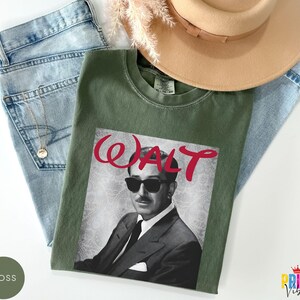 May include: An olive green t-shirt featuring a black and white image of a man wearing sunglasses, with the word "WALT" in red script. The shirt is laid on top of blue jeans and a tan hat.