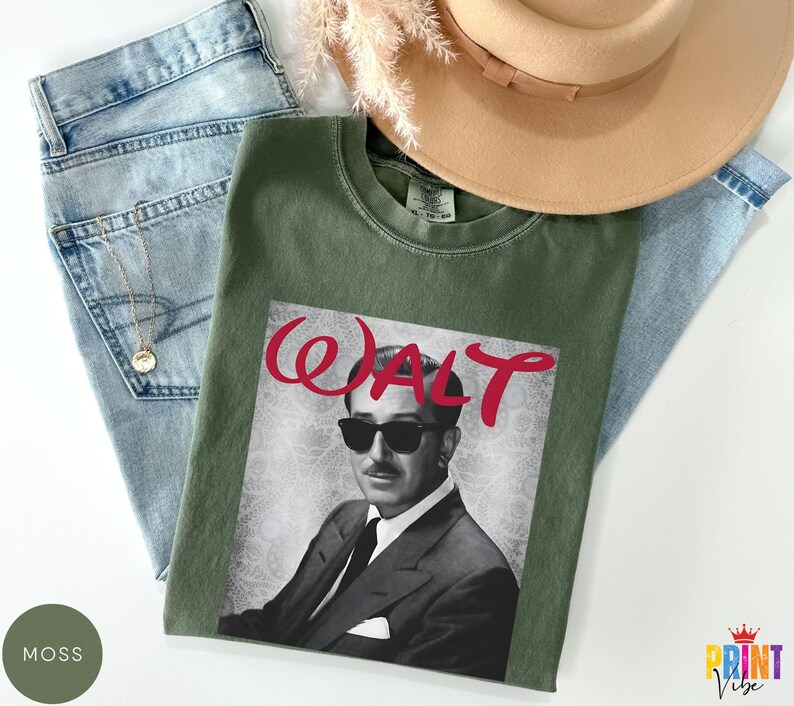May include: An olive green t-shirt featuring a black and white image of a man wearing sunglasses, with the word "WALT" in red script. The shirt is laid on top of blue jeans and a tan hat.