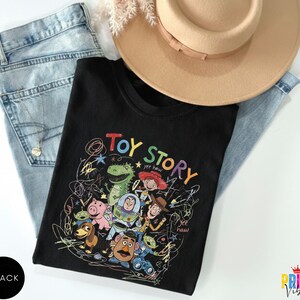 May include: A black t-shirt featuring a colorful graphic print of Toy Story characters including Woody, Buzz Lightyear, Rex, Jessie, Hamm, Slinky Dog, Mr. Potato Head, and Aliens, surrounded by scribbled lines and the text Toy Story and yee haw.