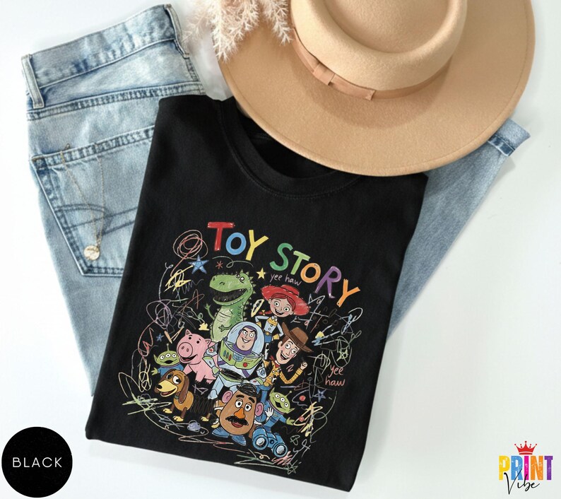 May include: A black t-shirt featuring a colorful graphic print of Toy Story characters including Woody, Buzz Lightyear, Rex, Jessie, Hamm, Slinky Dog, Mr. Potato Head, and Aliens, surrounded by scribbled lines and the text Toy Story and yee haw.