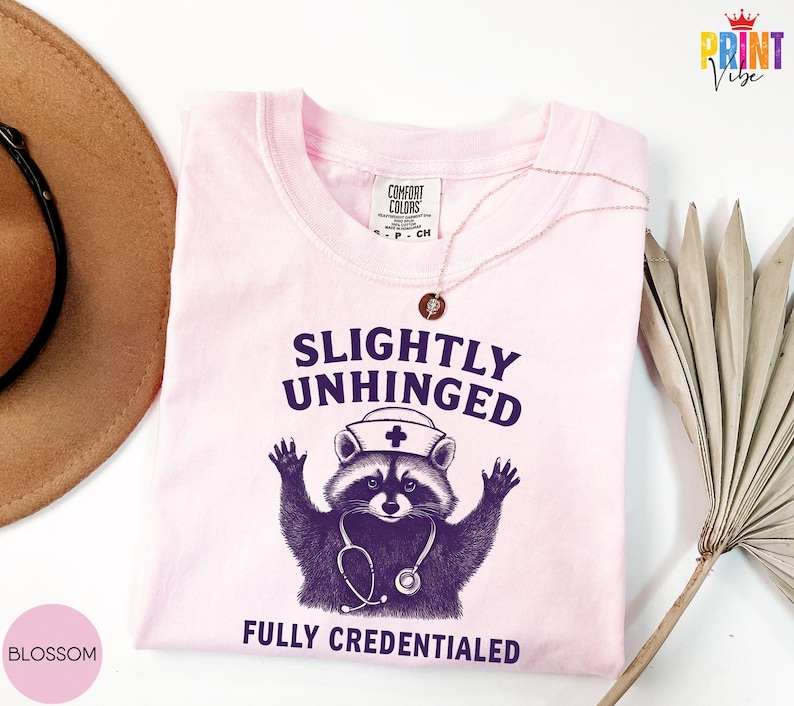 May include: A pale pink t-shirt featuring the text "SLIGHTLY UNHINGED" above a raccoon in a nurse's hat and stethoscope, with "FULLY CREDENTIALED" below. The shirt has a crew neck and short sleeves.