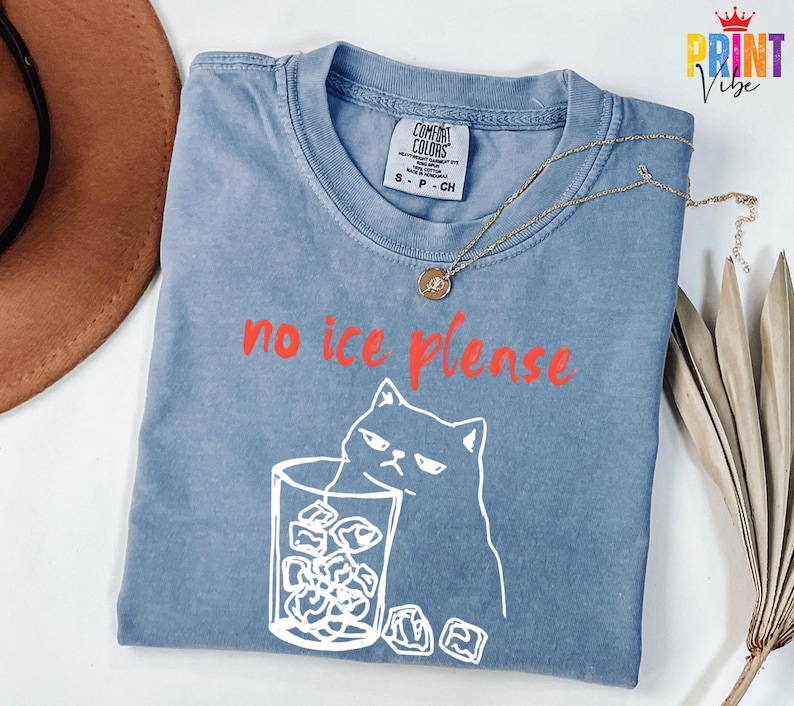 May include: A blue Comfort Colors t-shirt with the phrase "no ice please" in red. The shirt features a white graphic of a grumpy cat holding a glass of ice. A gold necklace with a pendant is on the shirt.