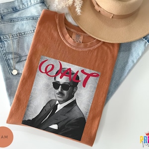May include: An orange t-shirt featuring a black and white image of a man wearing sunglasses, with the name "WALT" in red script. The shirt is laid on top of blue jeans and a tan hat.