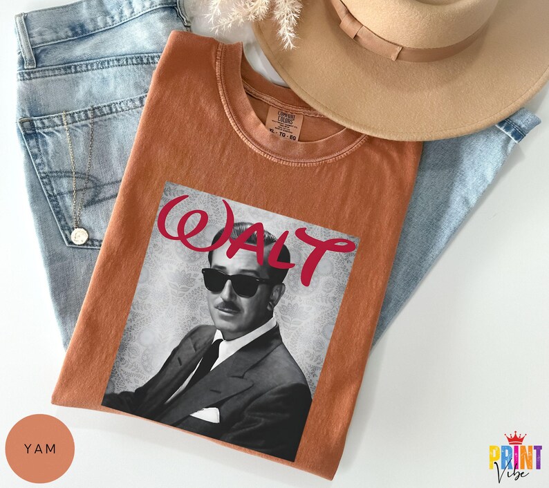 May include: An orange t-shirt featuring a black and white image of a man wearing sunglasses, with the name "WALT" in red script. The shirt is laid on top of blue jeans and a tan hat.