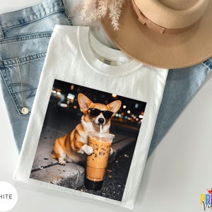 May include: White t-shirt featuring a color image of a corgi wearing sunglasses and holding an iced coffee. The corgi is brown and white. The t-shirt is laid on top of blue jeans and a tan hat.
