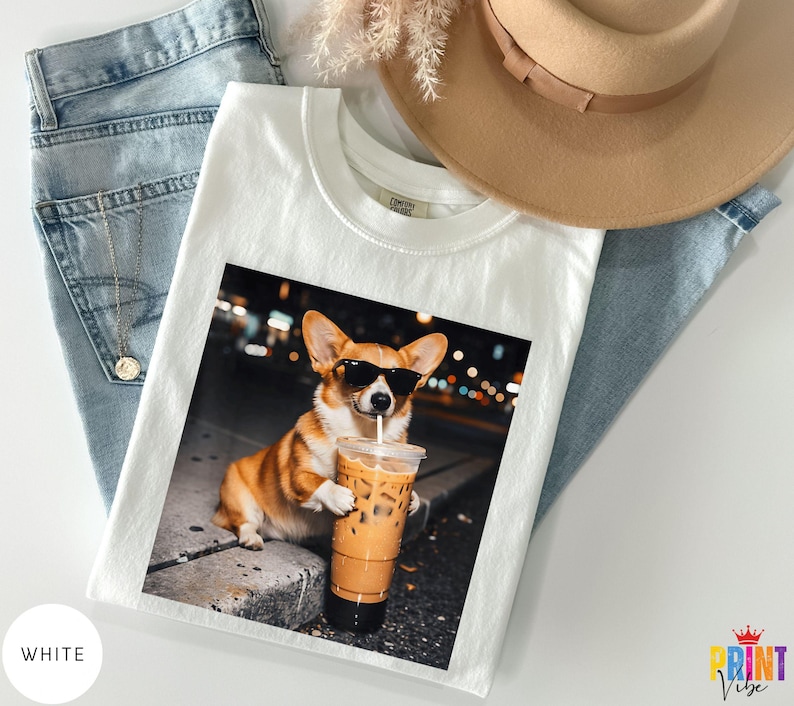 May include: White t-shirt featuring a color image of a corgi wearing sunglasses and holding an iced coffee. The corgi is brown and white. The t-shirt is laid on top of blue jeans and a tan hat.