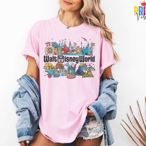 Puede incluir: Camiseta rosa claro con un gr&aacute;fico colorido de las atracciones de Walt Disney World. El dise&ntilde;o incluye una noria, un castillo y otros lugares emblem&aacute;ticos, con las palabras "Walt Disney World" en negrita.