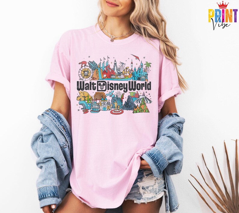 Puede incluir: Camiseta rosa claro con un gr&aacute;fico colorido de las atracciones de Walt Disney World. El dise&ntilde;o incluye una noria, un castillo y otros lugares emblem&aacute;ticos, con las palabras "Walt Disney World" en negrita.