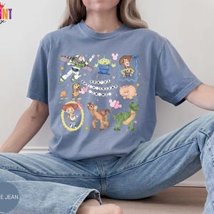 May include: A blue-gray short-sleeve t-shirt featuring a graphic print of various animated characters including a space ranger, a cowboy, a cowgirl, a dinosaur, and a pig. The text "YOU'VE GOT A FRIEND IN ME" is printed in the center with small stars.