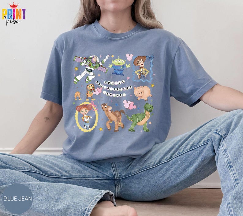 May include: A blue-gray short-sleeve t-shirt featuring a graphic print of various animated characters including a space ranger, a cowboy, a cowgirl, a dinosaur, and a pig. The text "YOU'VE GOT A FRIEND IN ME" is printed in the center with small stars.