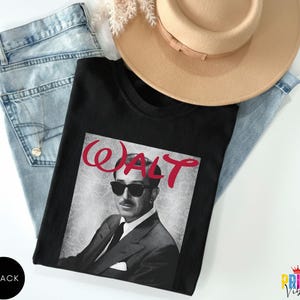 May include: Black crew neck t-shirt featuring a grayscale image of a man wearing sunglasses, with the name "WALT" in red script above. The shirt is laid on top of light blue jeans and next to a tan hat.