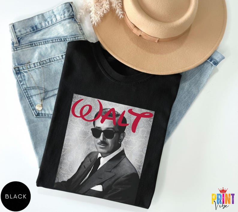 May include: Black crew neck t-shirt featuring a grayscale image of a man wearing sunglasses, with the name "WALT" in red script above. The shirt is laid on top of light blue jeans and next to a tan hat.