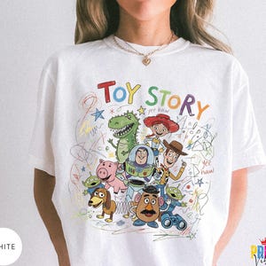 May include: A white t-shirt featuring a colorful graphic print of Toy Story characters including Woody, Buzz Lightyear, Jessie, Rex, Hamm, Slinky Dog, and the Aliens. The design includes the text Toy Story and yee haw with scribbled accents.