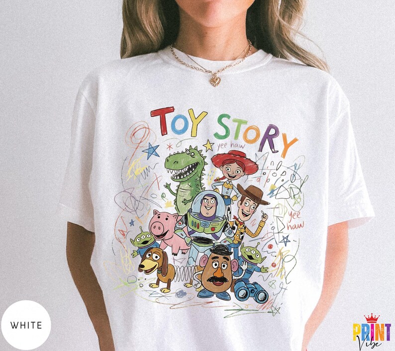 May include: A white t-shirt featuring a colorful graphic print of Toy Story characters including Woody, Buzz Lightyear, Jessie, Rex, Hamm, Slinky Dog, and the Aliens. The design includes the text Toy Story and yee haw with scribbled accents.