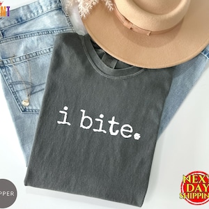 May include: A charcoal gray short-sleeve t-shirt with the white text i bite. printed on the front. The shirt is layered with a pair of light blue denim jeans, a tan wide-brimmed hat, and a thin gold chain necklace with a small circular pendant.