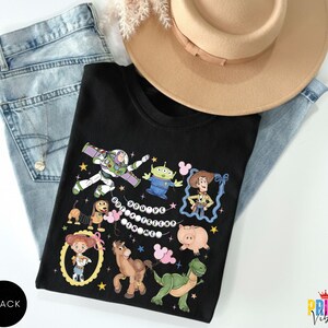 May include: A black t-shirt featuring a graphic print of various animated characters including a space ranger, a cowboy, a cowgirl, a green alien, a dinosaur, a pig, a horse, and a spring dog, with the text "YOU'VE GOT A FRIEND IN ME" in white.