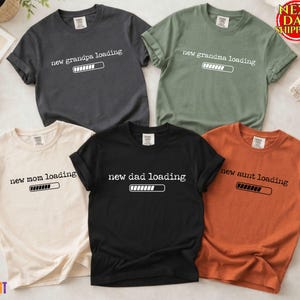 May include: Five short-sleeved t-shirts in various colors: charcoal, olive green, cream, black, and orange. Each shirt features the phrase "new grandpa loading," "new grandma loading," "new mom loading," "new dad loading," and "new aunt loading" with a loading bar graphic.