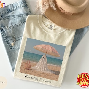 May include: An ivory t-shirt featuring a graphic print of a glittery white ghost standing on a sandy beach under a pink and white striped beach umbrella, with a straw tote bag nearby and the text Mentally, I'm here printed below the image.