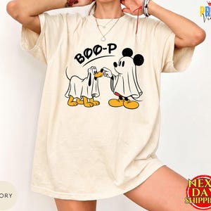 May include: An ivory short-sleeve t-shirt featuring a graphic of a dog and a mouse character, both wearing white ghost costumes. Above the characters, the text BOO-P is printed in black. The shirt has a crew neckline and a relaxed fit.